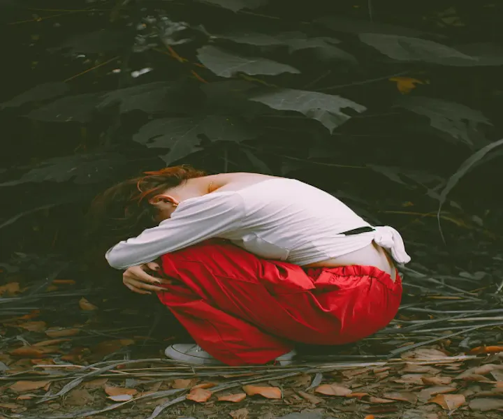 Gabor Maté’s H.E.A.L. Technique for Trauma Recovery. Image of a person wearing a white long-sleeve top and red pants crouches on the ground in a forest, head down and arms wrapped around their knees, surrounded by fallen leaves and dark green foliage, conveying a mood of sadness or introspection.