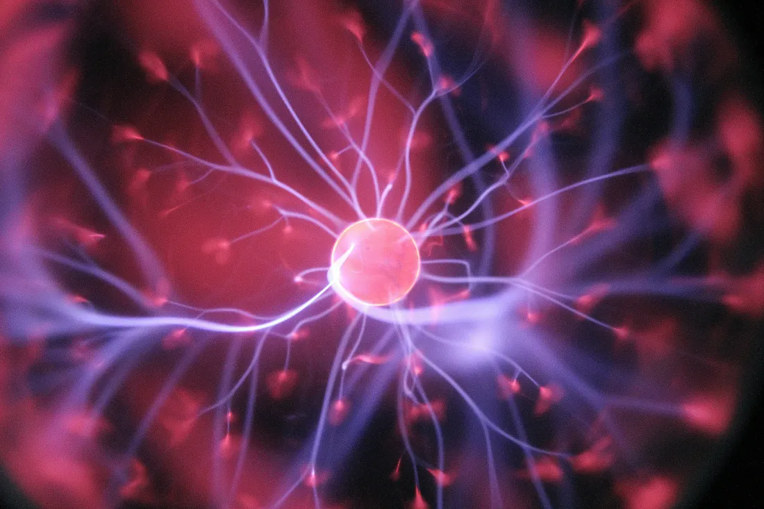 Recent neuroscience studies suggest the brain predicts trauma. Abstract close-up of glowing purple and pink electrical tendrils radiating from a bright central orb, resembling neurons or brain signals in a dark background.